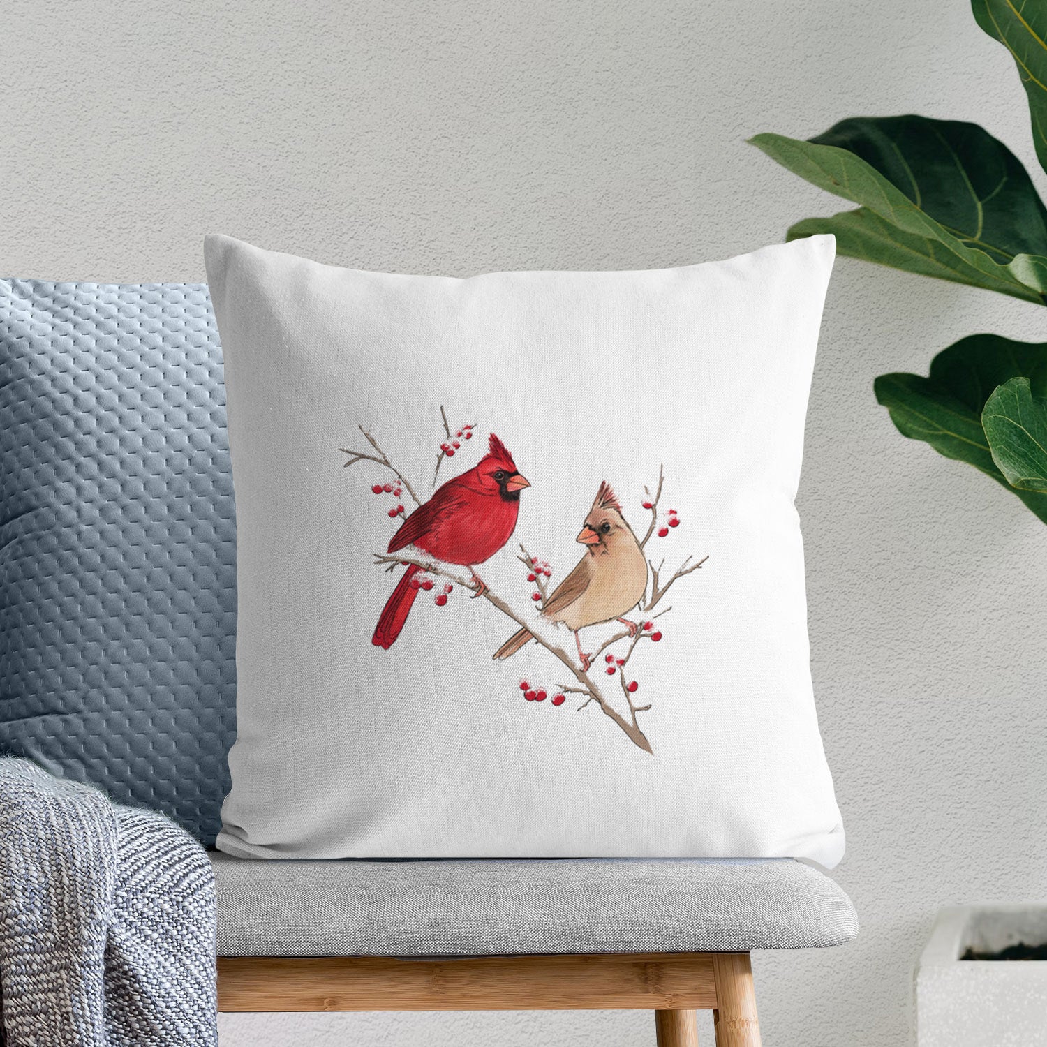 White pillow cover printed with a full-color illustration of a male and female northern cardinal perched on a snowy branch with red berries, on a bench beside plants.