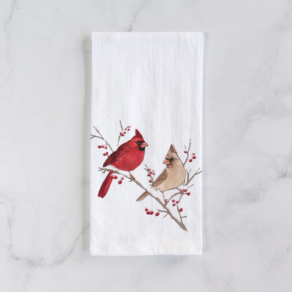 White cotton tea towel laid flat on marble, printed with a full-color illustration of a male and female northern cardinal perched on a snowy branch with red berries.