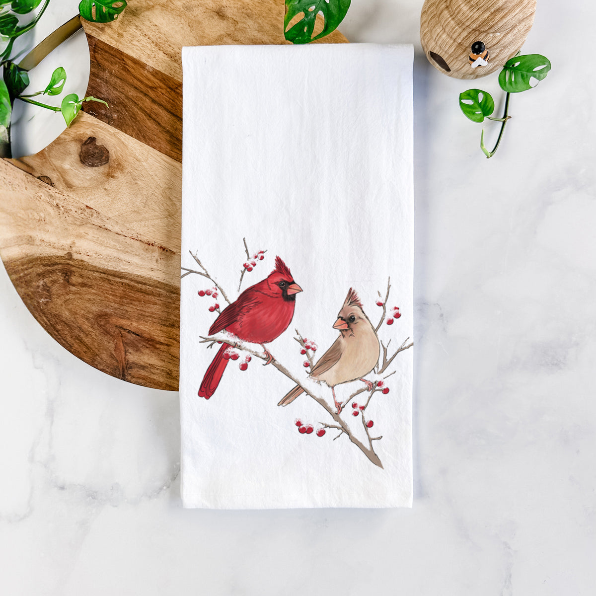 White cotton tea towel printed with a full-color illustration of a male and female northern cardinal perched on a snowy branch with red berries, on a marble counter beside a round wooden cutting board and greenery.