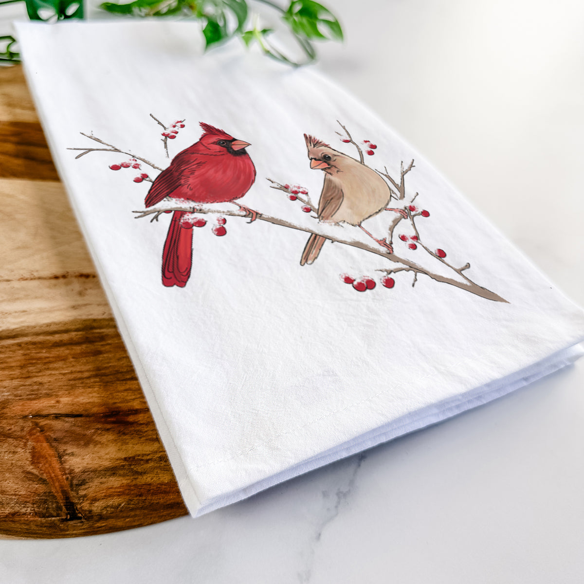 Close-up of a folded white tea towel on a wooden board, showing a full-color illustration of a male and female northern cardinal perched on a snowy branch with red berries.