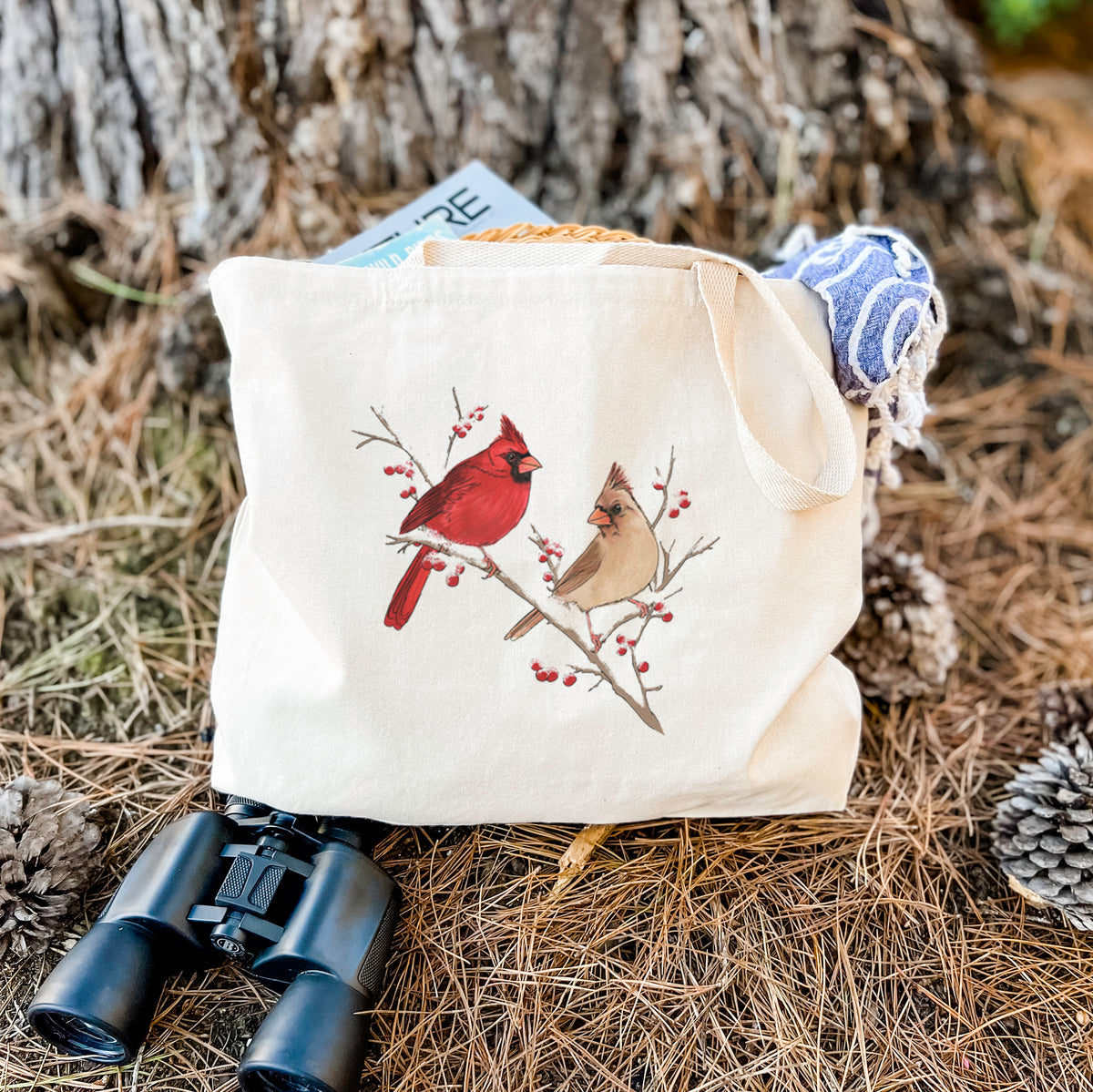 Canvas tote bag printed with a full-color illustration of a male and female northern cardinal perched on a snowy branch with red berries, resting on dry grass beside a pair of binoculars.