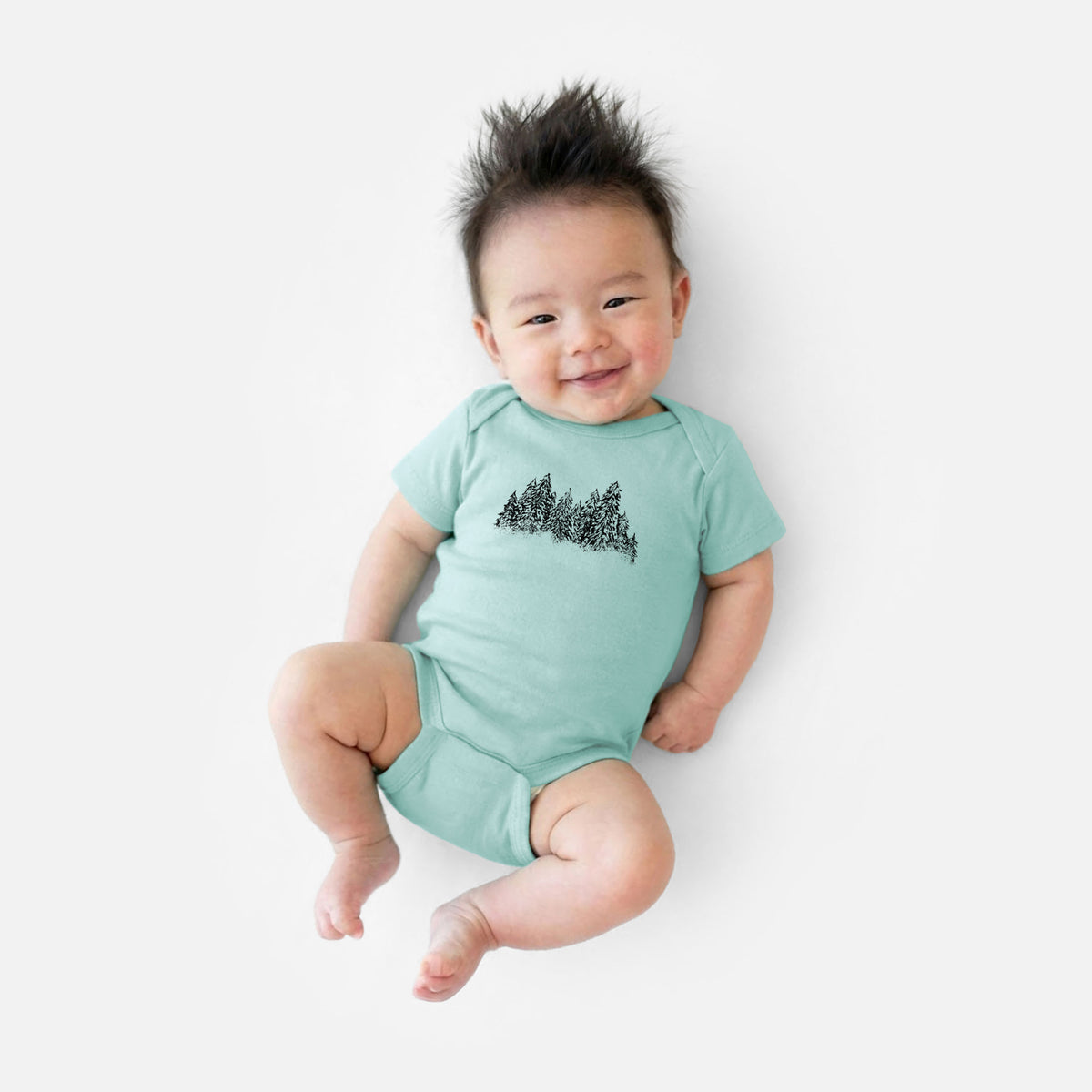 Smiling baby boy wearing a saltwater baby bodysuit printed with a black ink illustration of a row of snow-dusted evergreen trees.