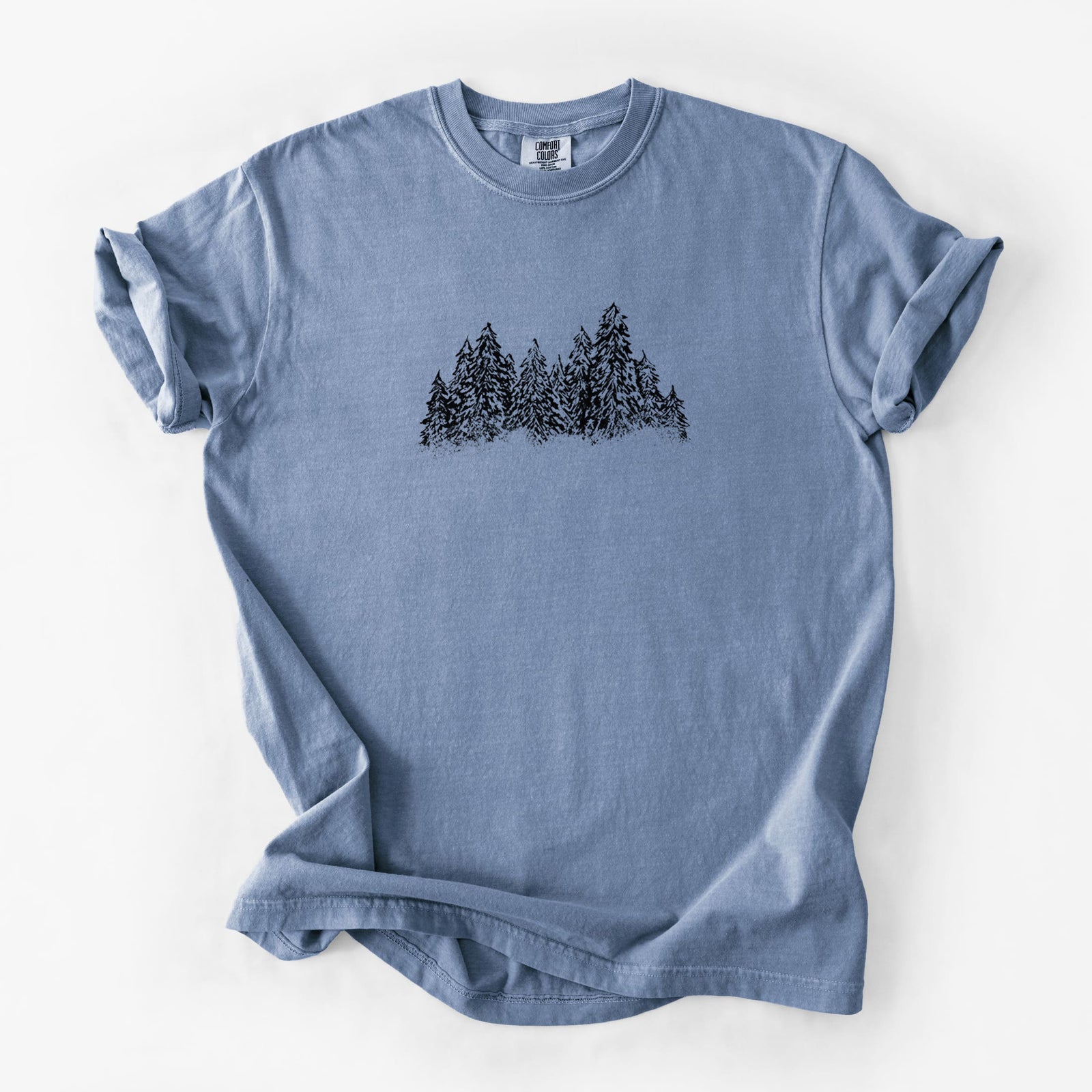 Blue jean Comfort Colors t-shirt laid flat, printed with a black ink illustration of a row of snow-dusted evergreen trees.