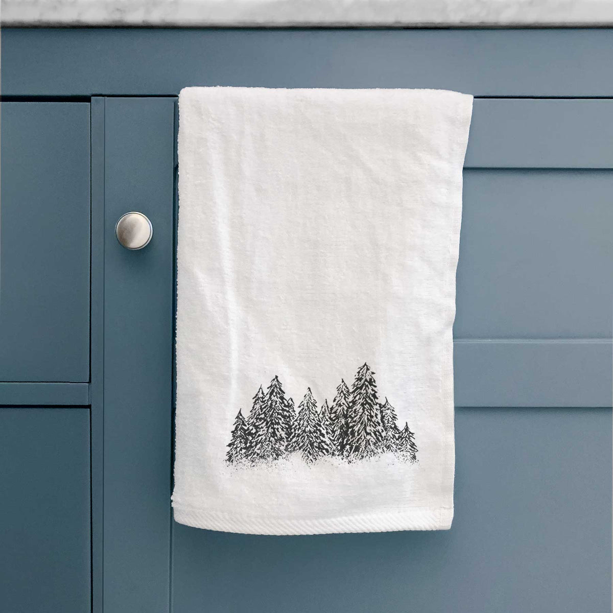 White hand towel printed with a black ink illustration of a row of snow-dusted evergreen trees, hanging on a cabinet door.