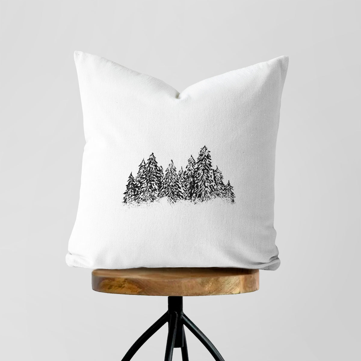 White pillow cover printed with a black ink illustration of a row of snow-dusted evergreen trees, on a wooden stool.