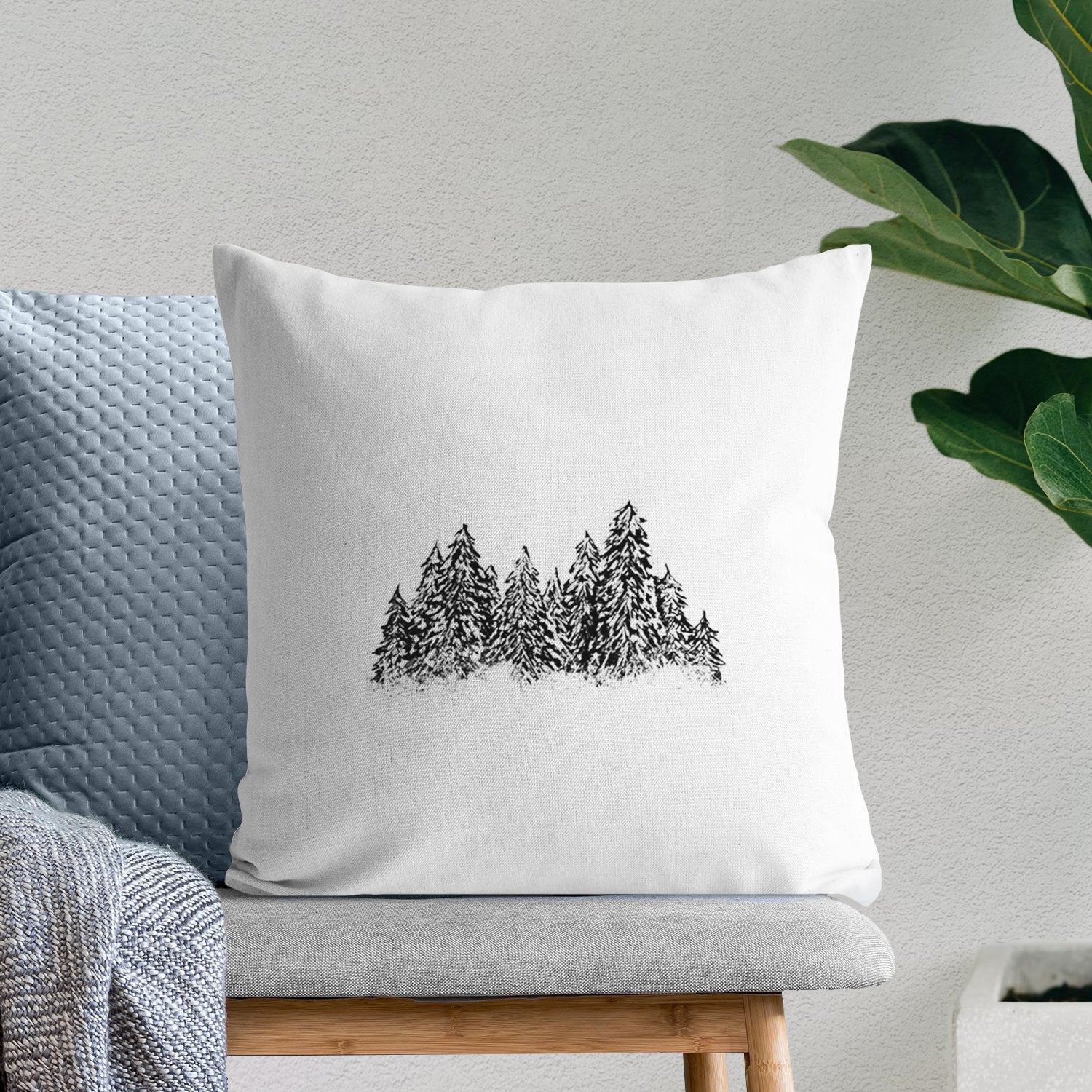 White pillow cover printed with a black ink illustration of a row of snow-dusted evergreen trees, on a bench beside plants.