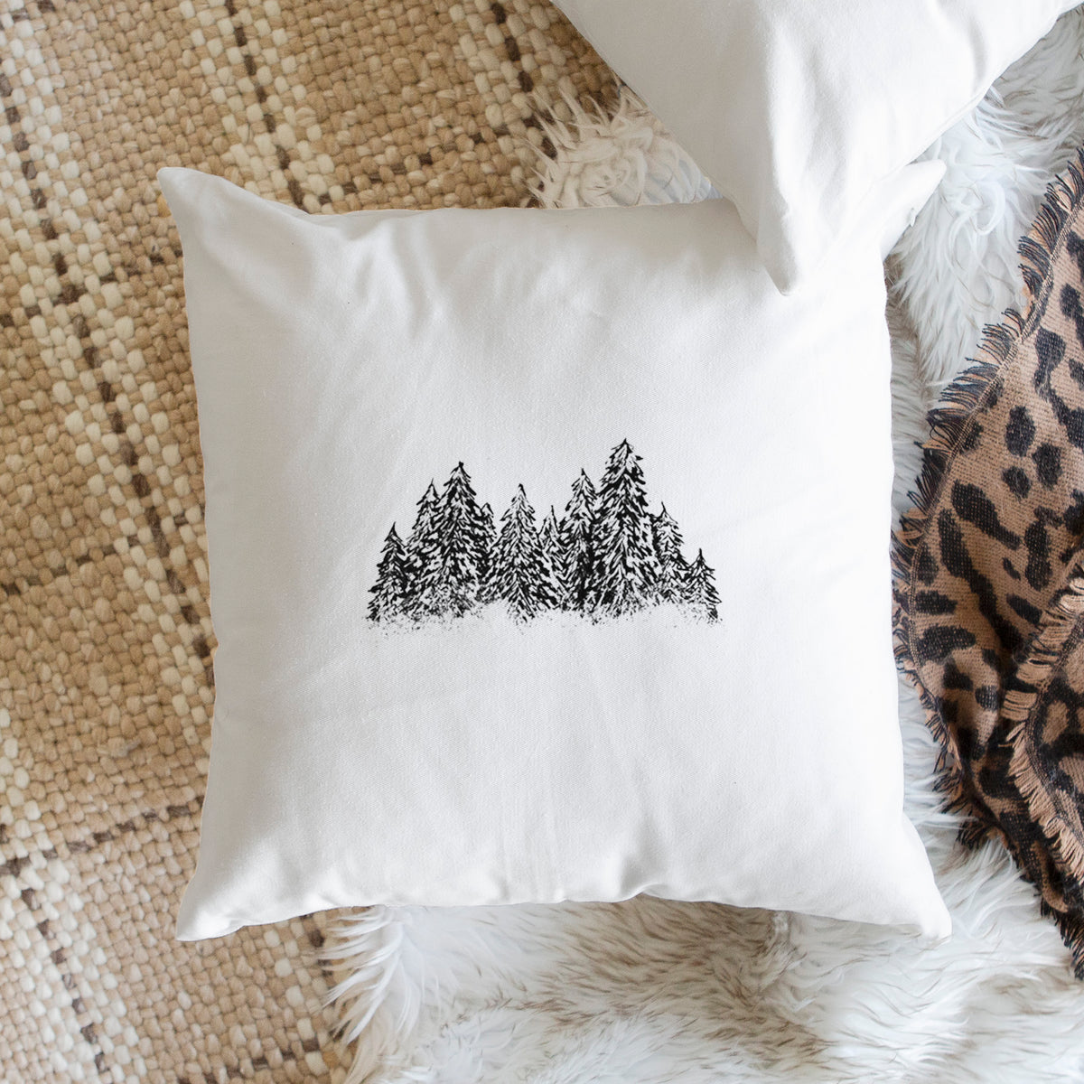 White pillow cover printed with a black ink illustration of a row of snow-dusted evergreen trees, on a woven chair.