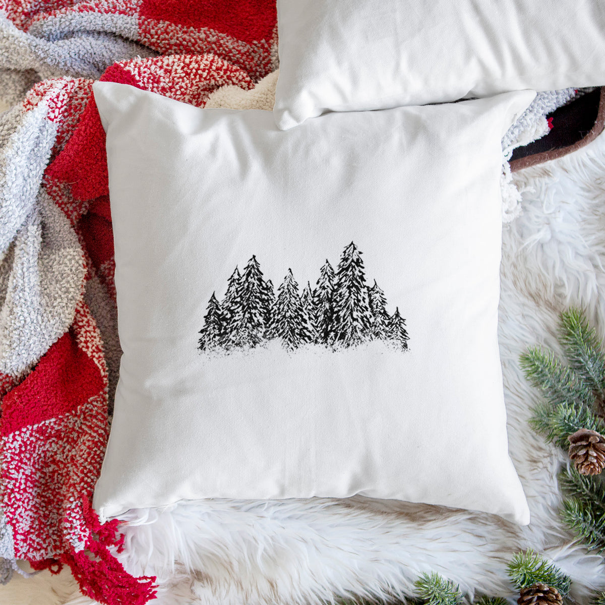 White pillow cover printed with a black ink illustration of a row of snow-dusted evergreen trees, on a woven chair.