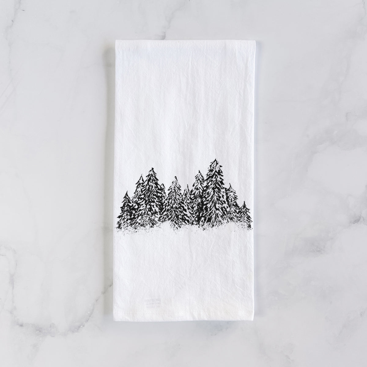 White cotton tea towel laid flat on marble, printed with a black ink illustration of a row of snow-dusted evergreen trees.