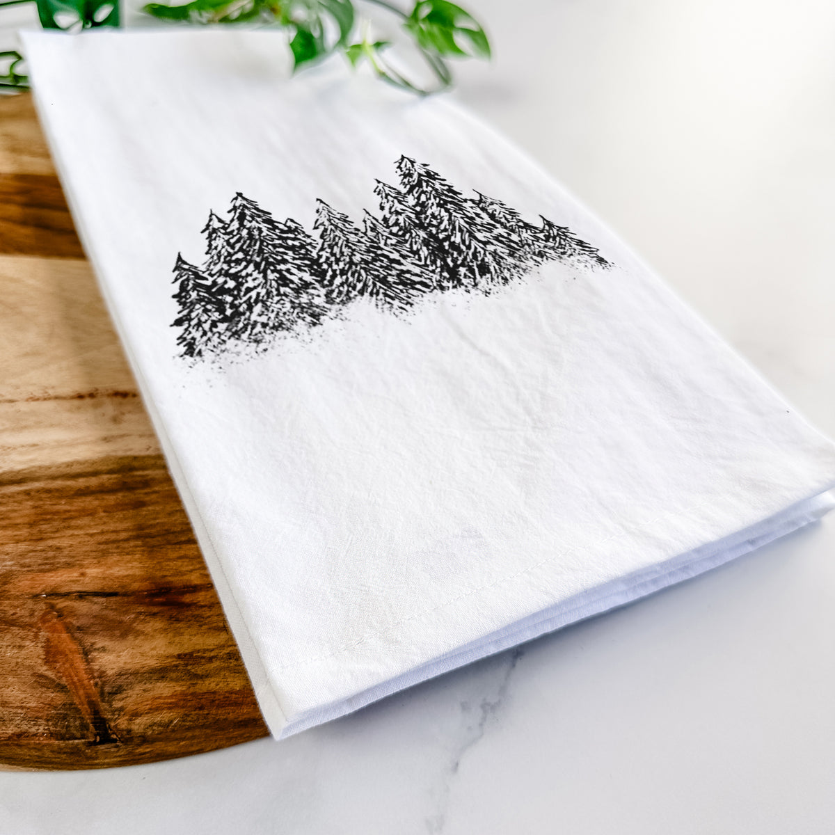 Close-up of a folded white tea towel on a wooden board, showing a black ink illustration of a row of snow-dusted evergreen trees.