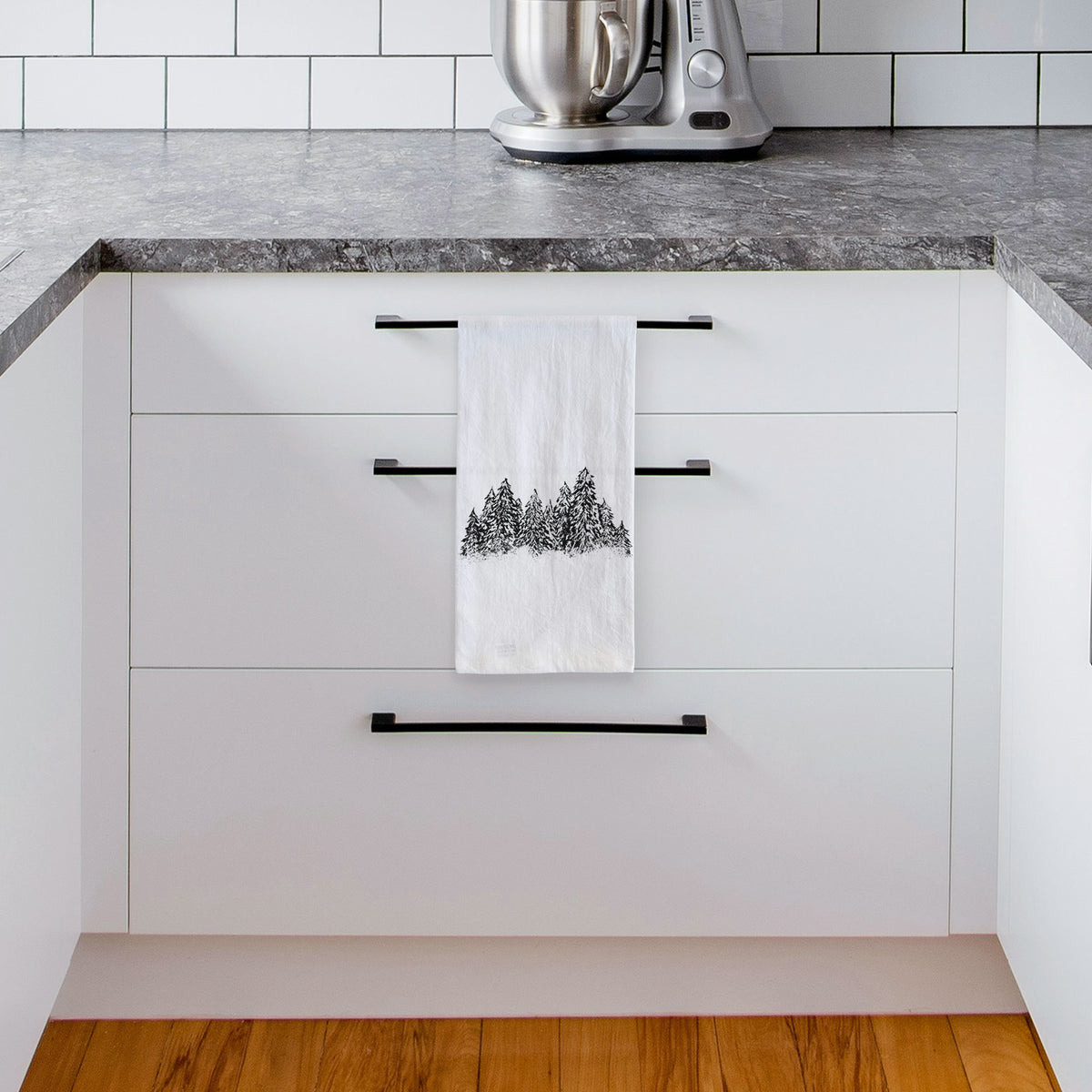 White tea towel printed with a black ink illustration of a row of snow-dusted evergreen trees, hanging from a drawer handle in a bright white kitchen.