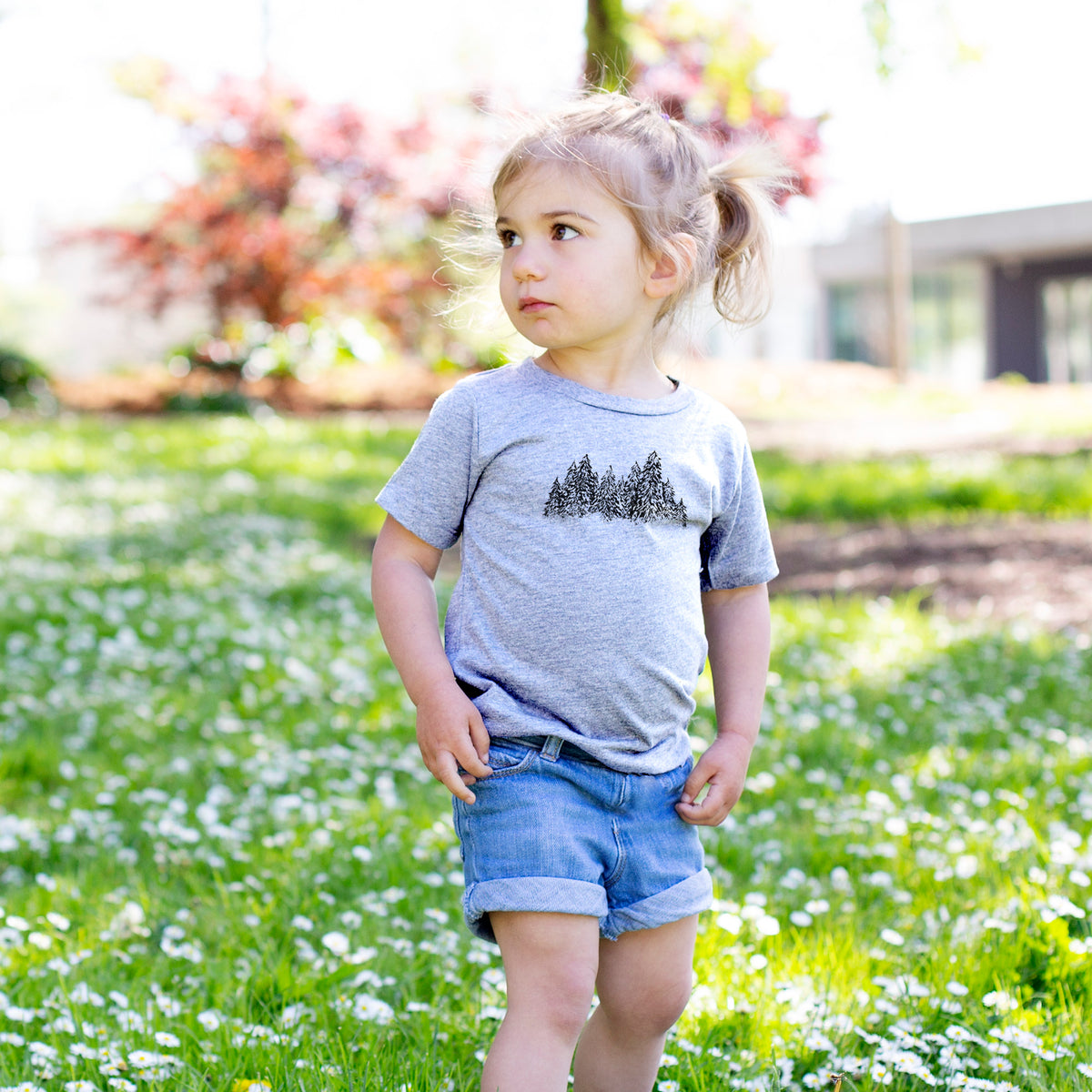 Toddler girl walking outdoors wearing a t-shirt printed with a black ink illustration of a row of snow-dusted evergreen trees.