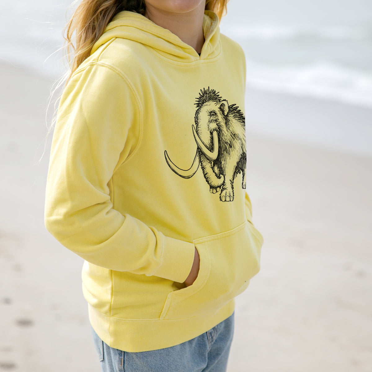 Girl wearing a youth pigment dyed hoodie printed with a black ink illustration of a woolly mammoth with long curved tusks.
