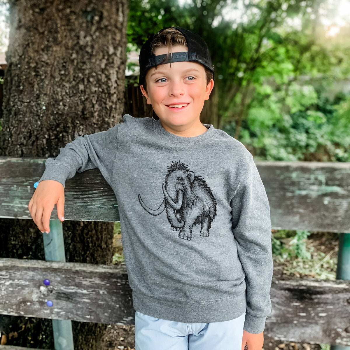 Boy wearing a youth crewneck sweatshirt printed with a black ink illustration of a woolly mammoth with long curved tusks, outdoors.