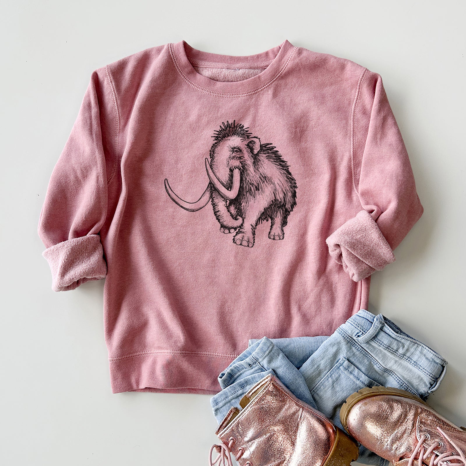 Boy wearing a youth crewneck sweatshirt printed with a black ink illustration of a woolly mammoth with long curved tusks, outdoors.