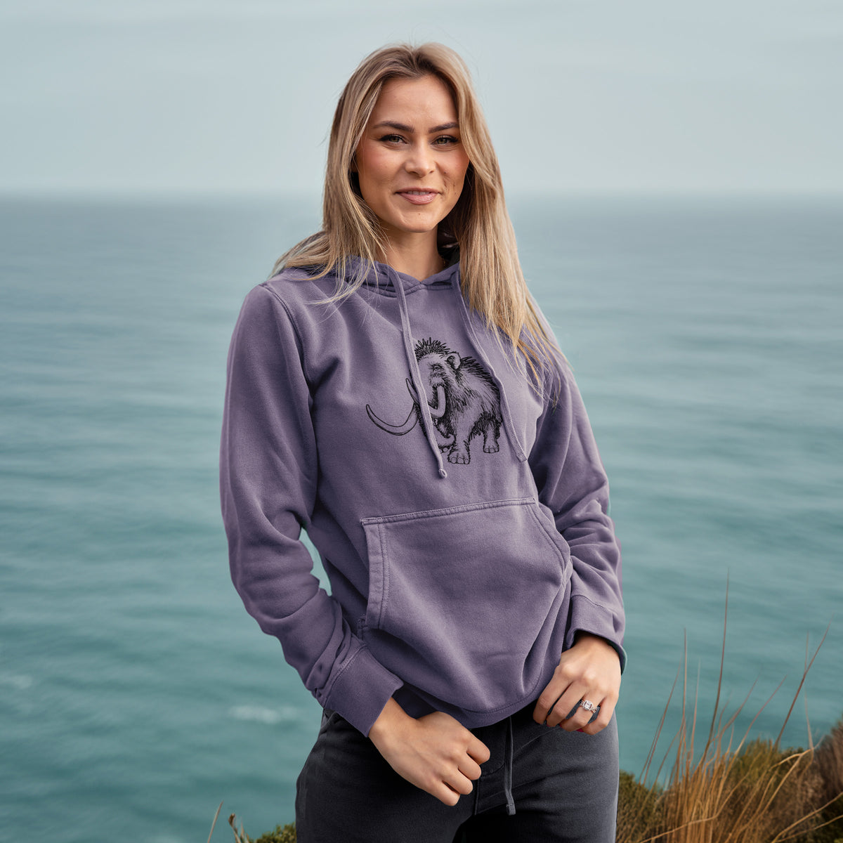 Woman wearing a plum purple pigment dyed hoodie printed with a black ink illustration of a woolly mammoth with long curved tusks.