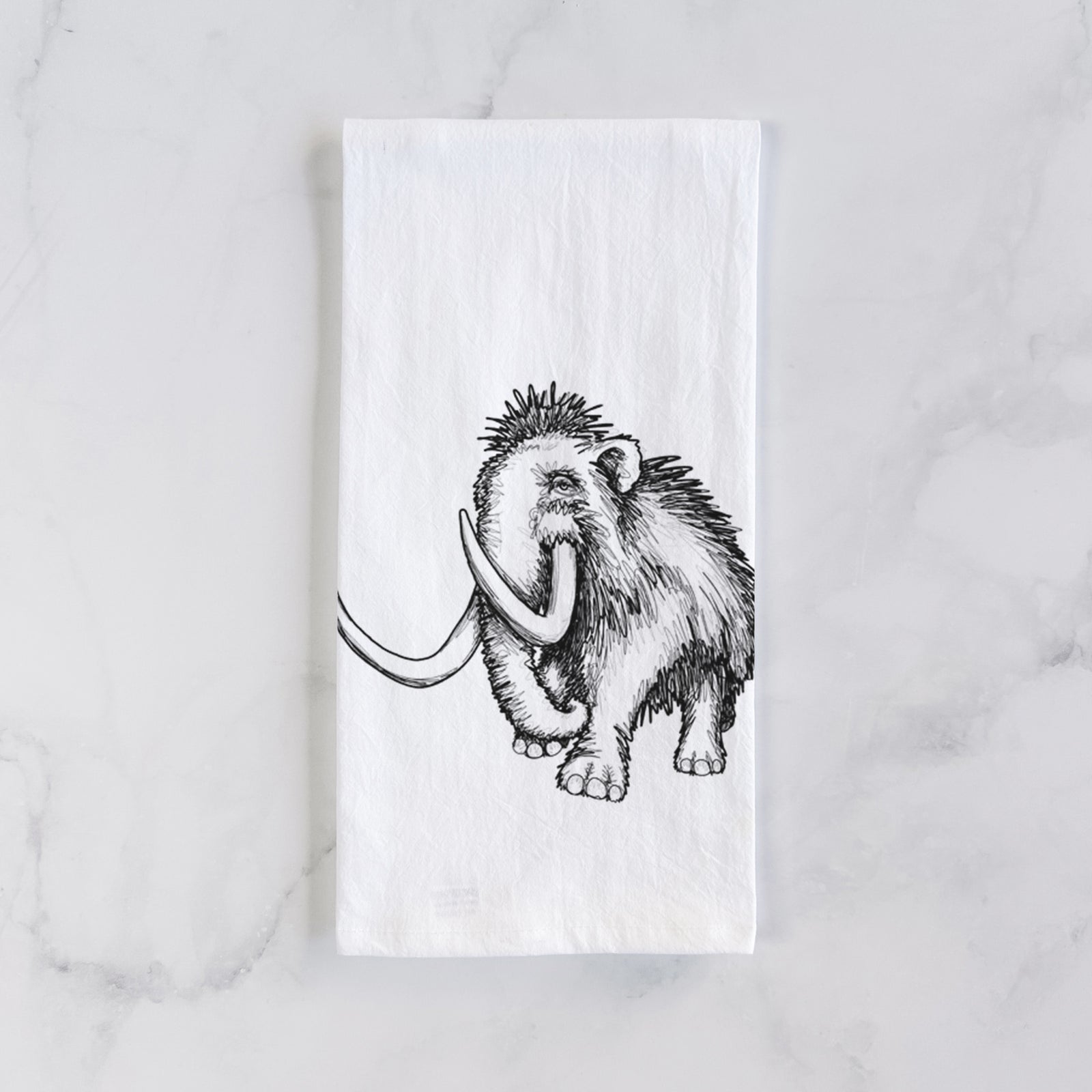 White cotton tea towel printed with a black ink illustration of a woolly mammoth with long curved tusks, on a marble counter beside a round wooden cutting board and greenery.