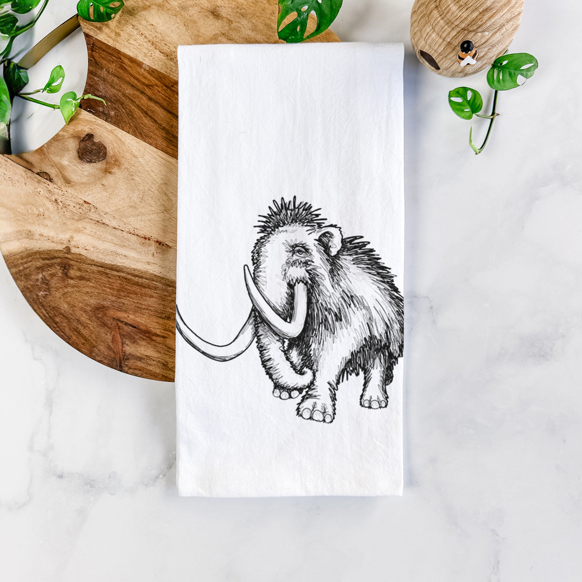 White cotton tea towel printed with a black ink illustration of a woolly mammoth with long curved tusks, on a marble counter beside a round wooden cutting board and greenery.