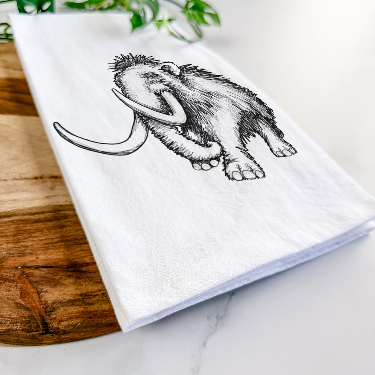 Close-up of a folded white tea towel on a wooden board, showing a black ink illustration of a woolly mammoth with long curved tusks.