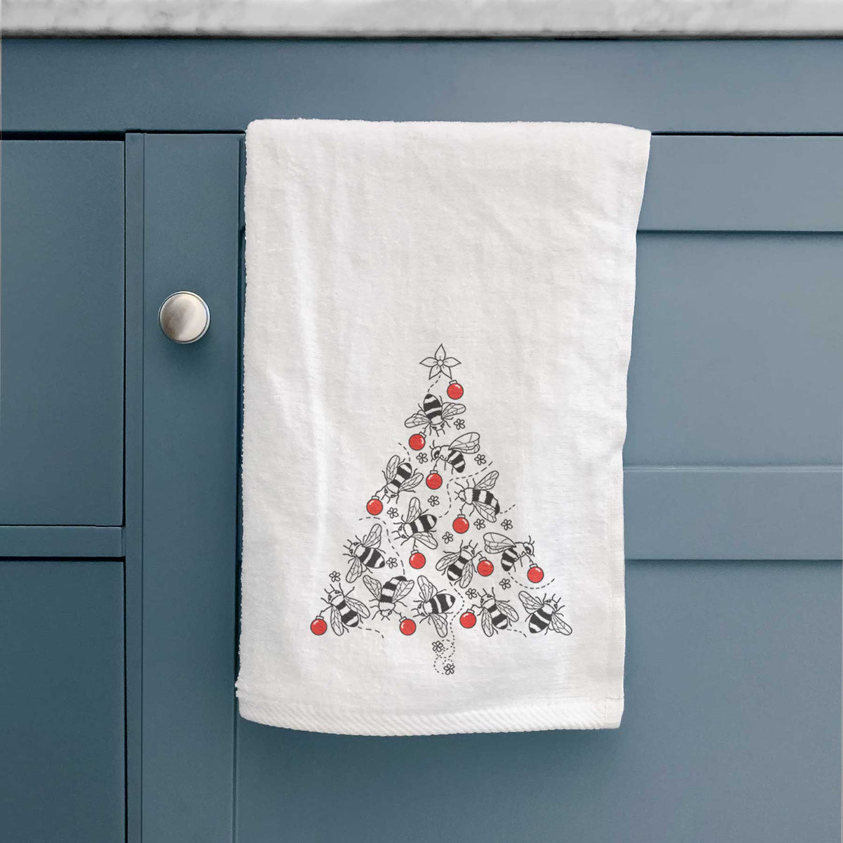 White hand towel printed with a Christmas tree made of black ink bees with red ornaments and a star on top, hanging on a cabinet door.