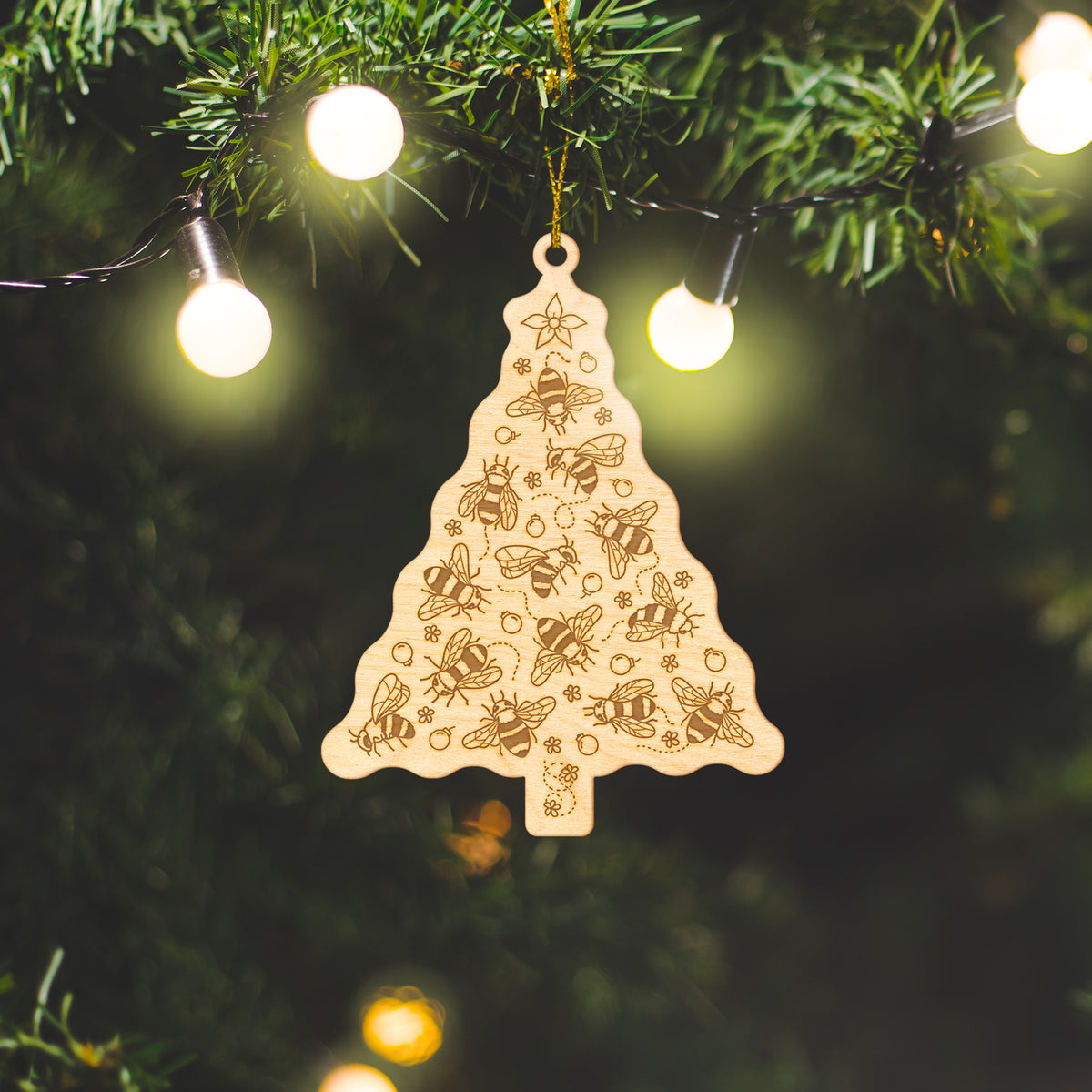 Wooden ornament featuring a Christmas tree made of engraved bees with red ornaments and a star on top, on a knit blanket with pine cones.