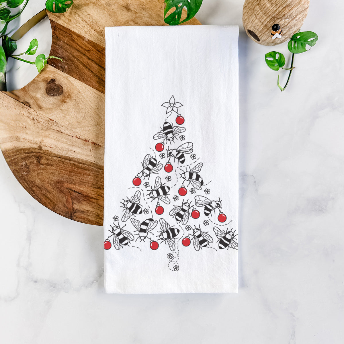 White cotton tea towel printed with a Christmas tree made of black ink bees with red ornaments and a star on top, on a marble counter beside a round wooden cutting board and greenery.