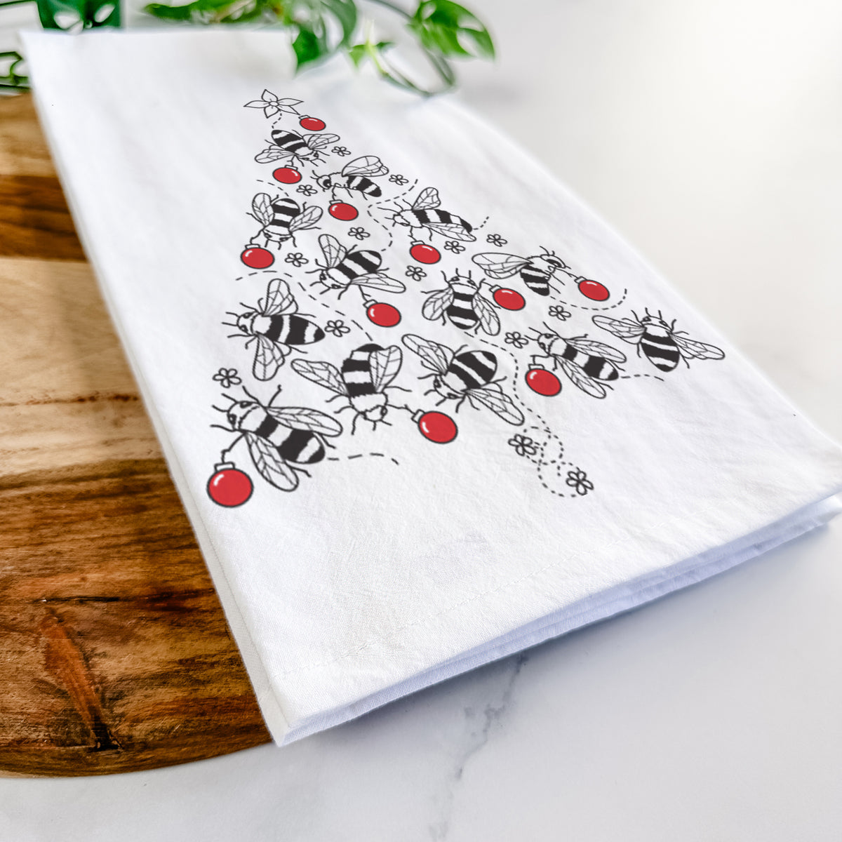 Close-up of a folded white tea towel on a wooden board, showing a Christmas tree made of black ink bees with red ornaments and a star on top.