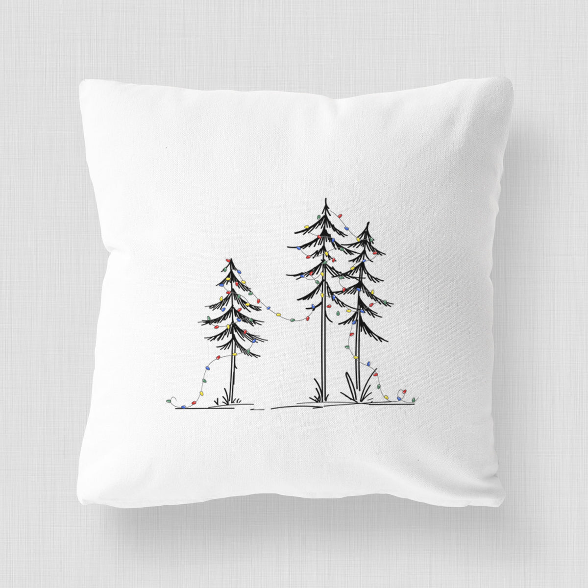 White pillow cover printed with three slender black ink pine trees strung with a garland of multicolored holiday lights.
