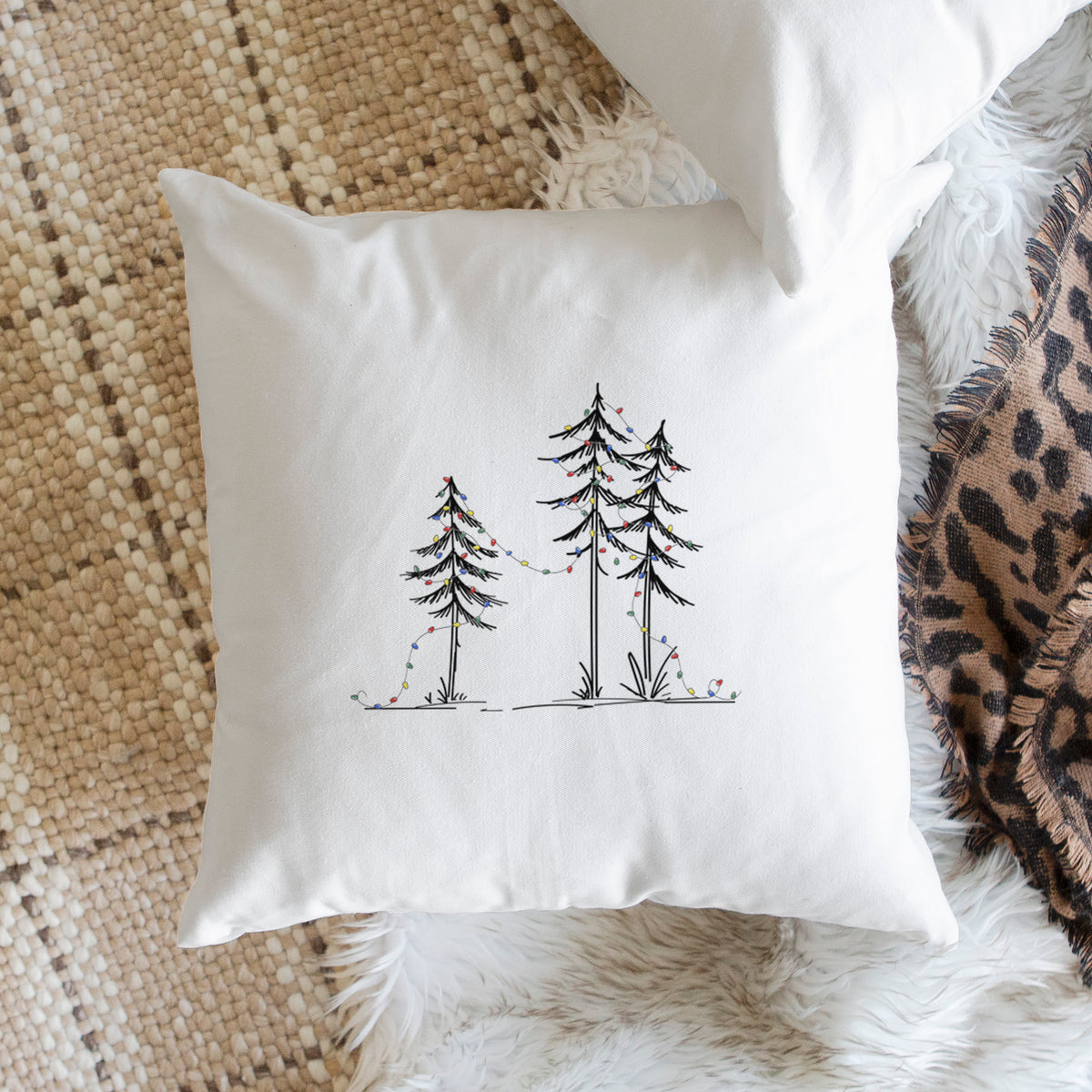 White pillow cover printed with three slender black ink pine trees strung with a garland of multicolored holiday lights, on a woven chair.