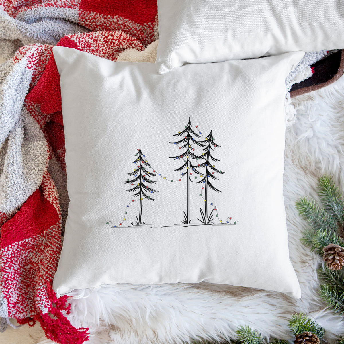 White pillow cover printed with three slender black ink pine trees strung with a garland of multicolored holiday lights, on a woven chair.