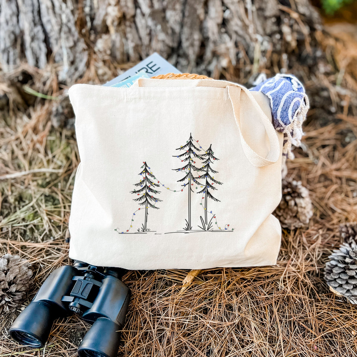 Canvas tote bag printed with three slender black ink pine trees strung with a garland of multicolored holiday lights, resting on dry grass beside a pair of binoculars.