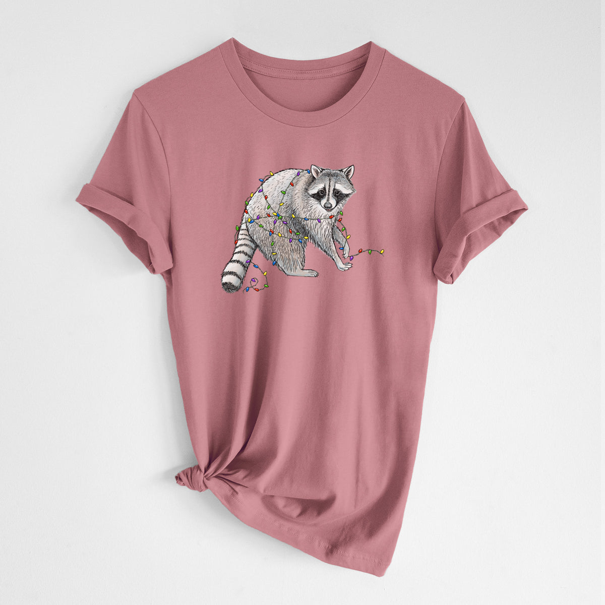 Mauve pink lightweight cotton t-shirt laid flat with the sleeves rolled, printed with a black ink raccoon tangled in a string of colorful holiday lights.