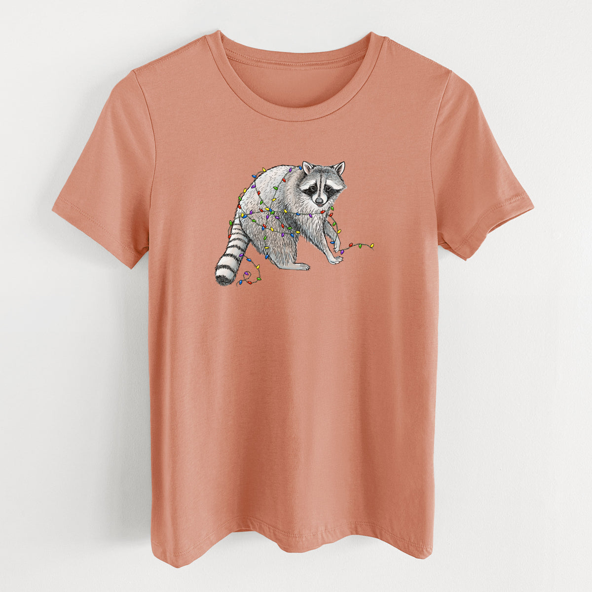 Terracotta orange women's relaxed fit cotton t-shirt laid flat, printed with a black ink raccoon tangled in a string of colorful holiday lights.