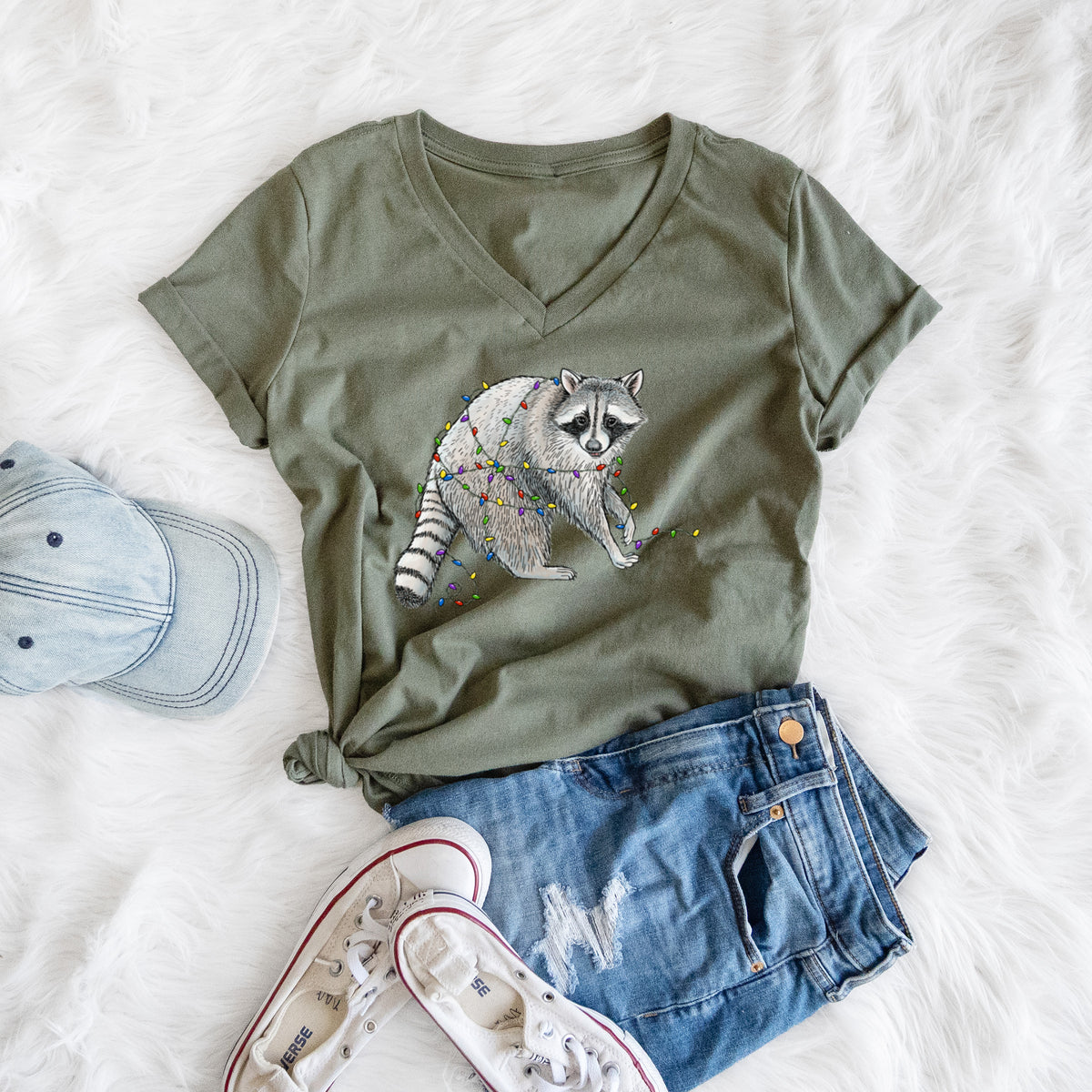Military green women's relaxed fit cotton V-neck t-shirt printed with a black ink raccoon tangled in a string of colorful holiday lights, styled flat on a white faux fur rug with a cap, jeans and sneakers.