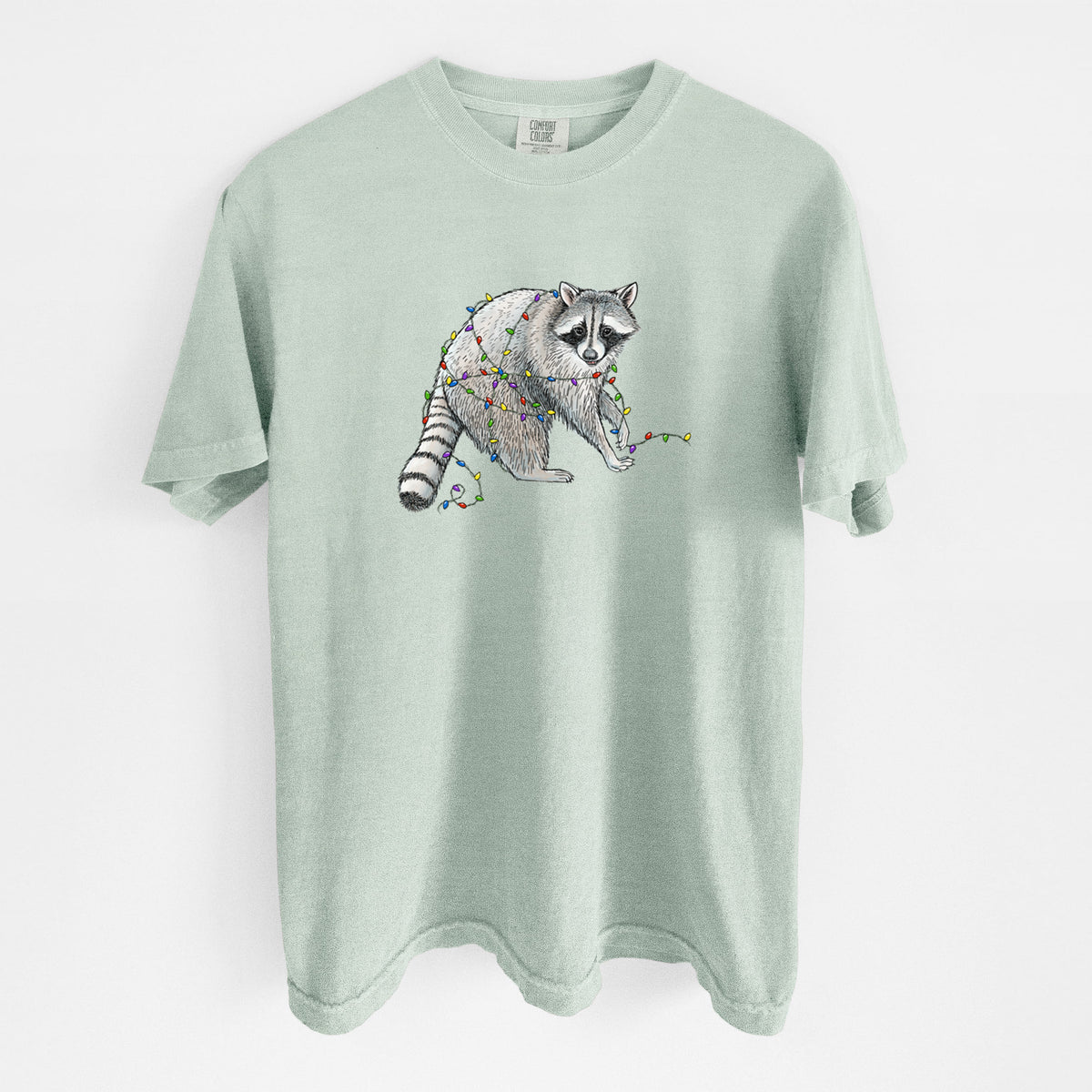 Bay green Comfort Colors t-shirt laid flat, printed with a black ink raccoon tangled in a string of colorful holiday lights.