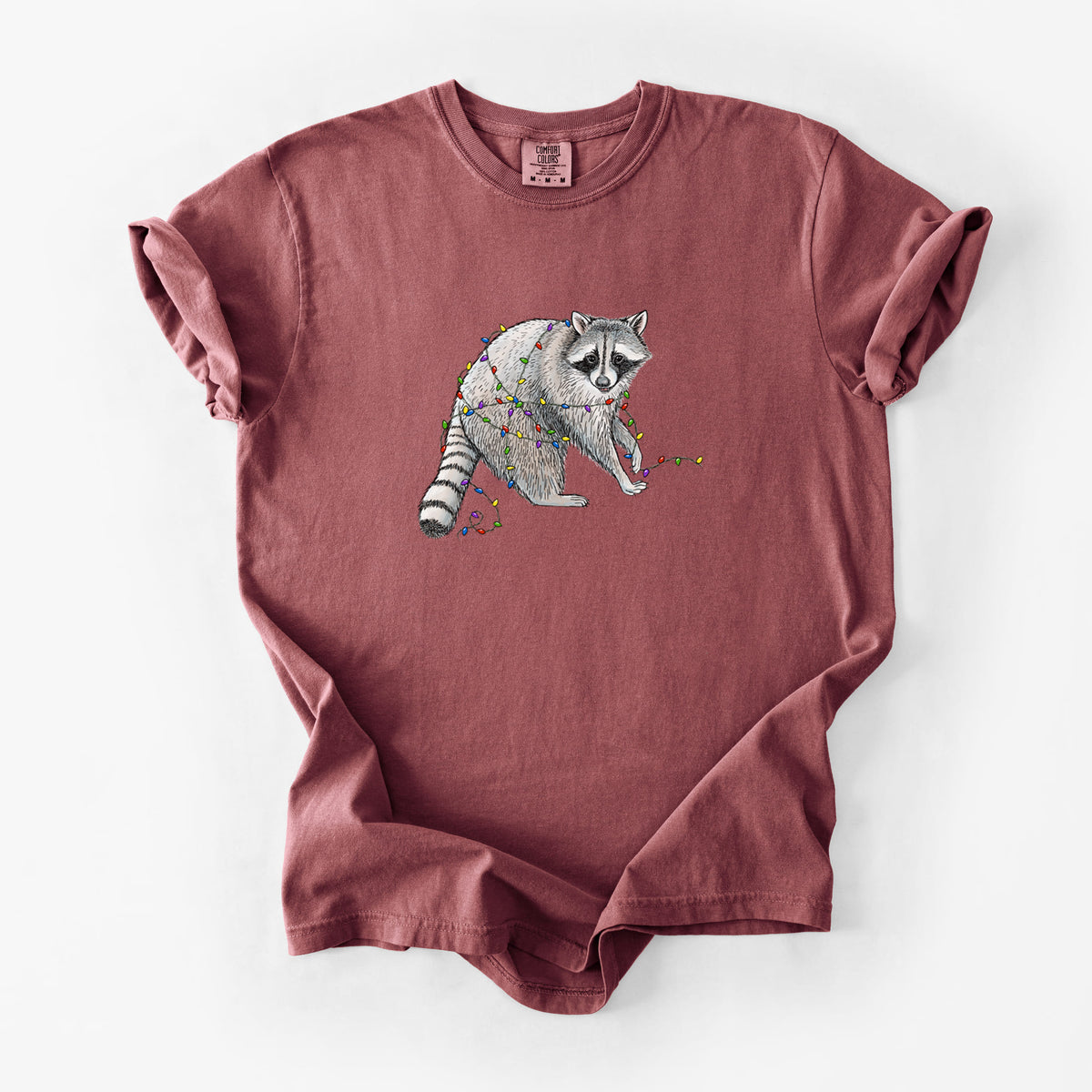 Brick red Comfort Colors t-shirt laid flat, printed with a black ink raccoon tangled in a string of colorful holiday lights.