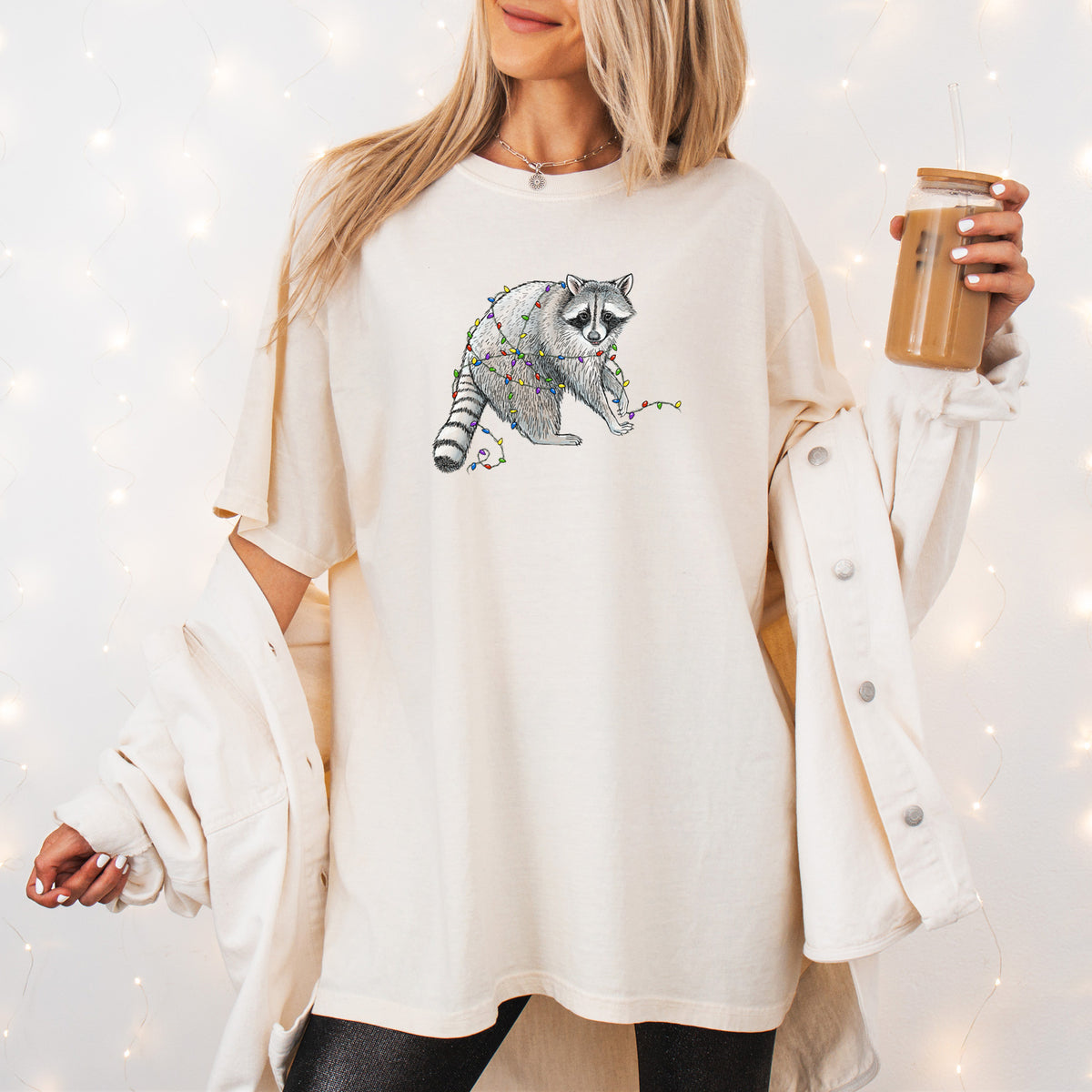 Woman wearing a Comfort Colors t-shirt printed with a black ink raccoon tangled in a string of colorful holiday lights, outdoors.