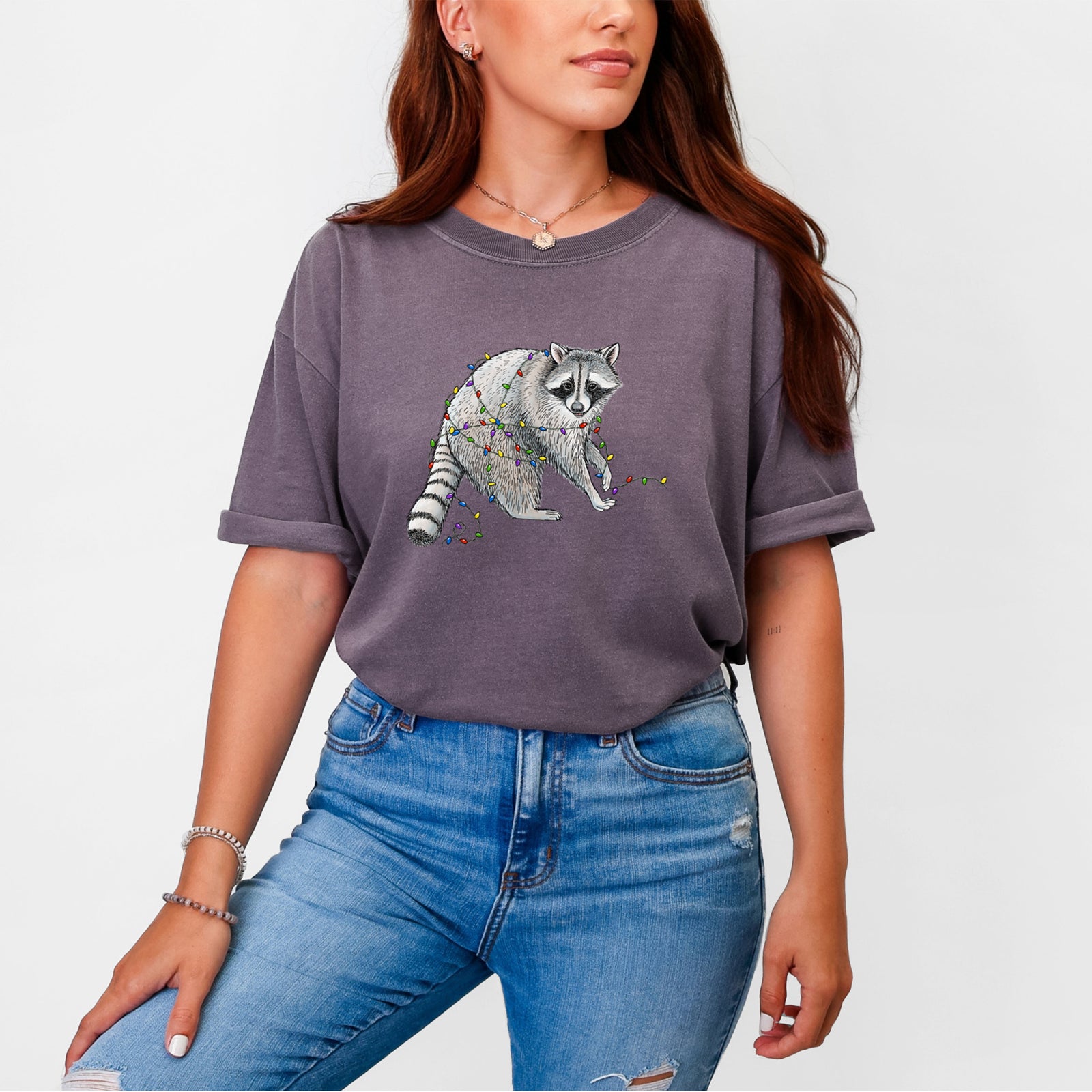 Blue jean Comfort Colors t-shirt laid flat, printed with a black ink raccoon tangled in a string of colorful holiday lights.