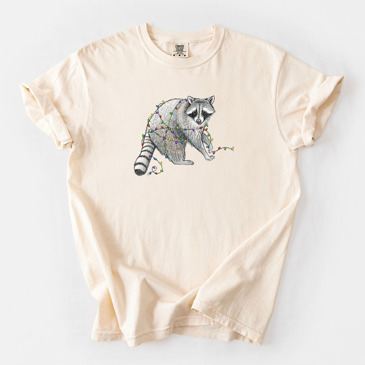Ivory Comfort Colors t-shirt laid flat, printed with a black ink raccoon tangled in a string of colorful holiday lights.