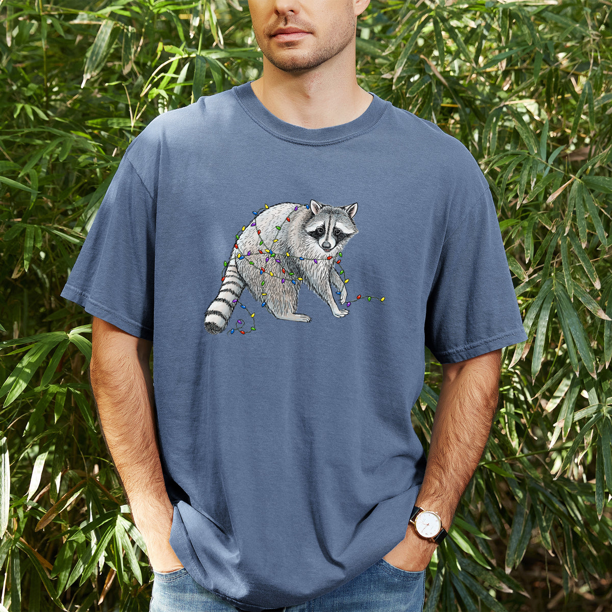 Man wearing a Comfort Colors t-shirt printed with a black ink raccoon tangled in a string of colorful holiday lights, outdoors.
