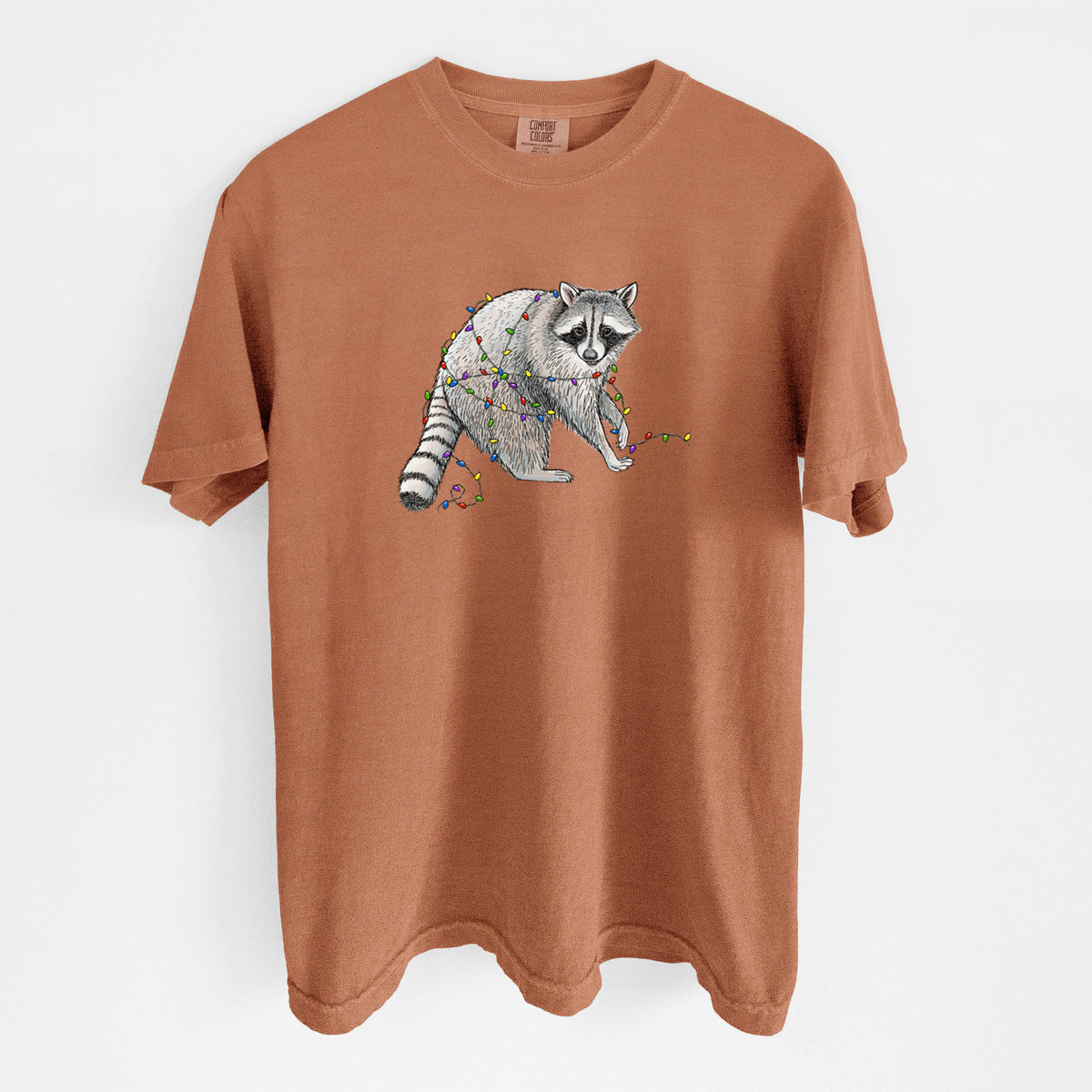 Yam burnt orange Comfort Colors t-shirt laid flat, printed with a black ink raccoon tangled in a string of colorful holiday lights.