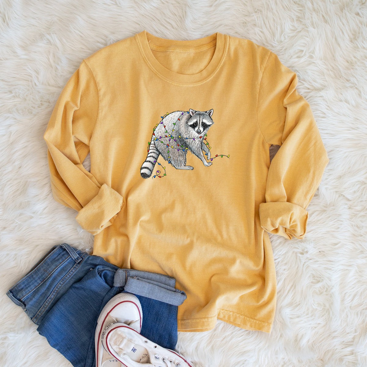 Golden wheat yellow Comfort Colors long sleeve shirt printed with a black ink raccoon tangled in a string of colorful holiday lights, shown in a lifestyle photo.