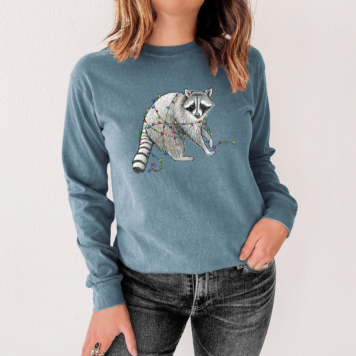 Steel blue Comfort Colors long sleeve shirt printed with a black ink raccoon tangled in a string of colorful holiday lights, shown in a lifestyle photo.