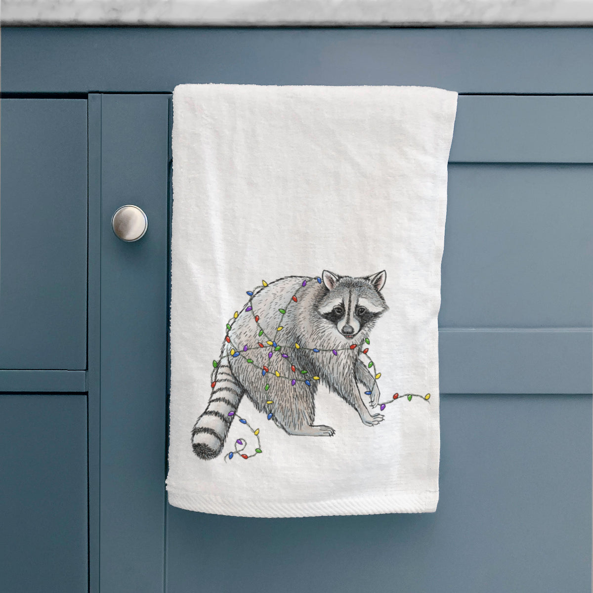 White hand towel printed with a black ink raccoon tangled in a string of colorful holiday lights, hanging on a cabinet door.