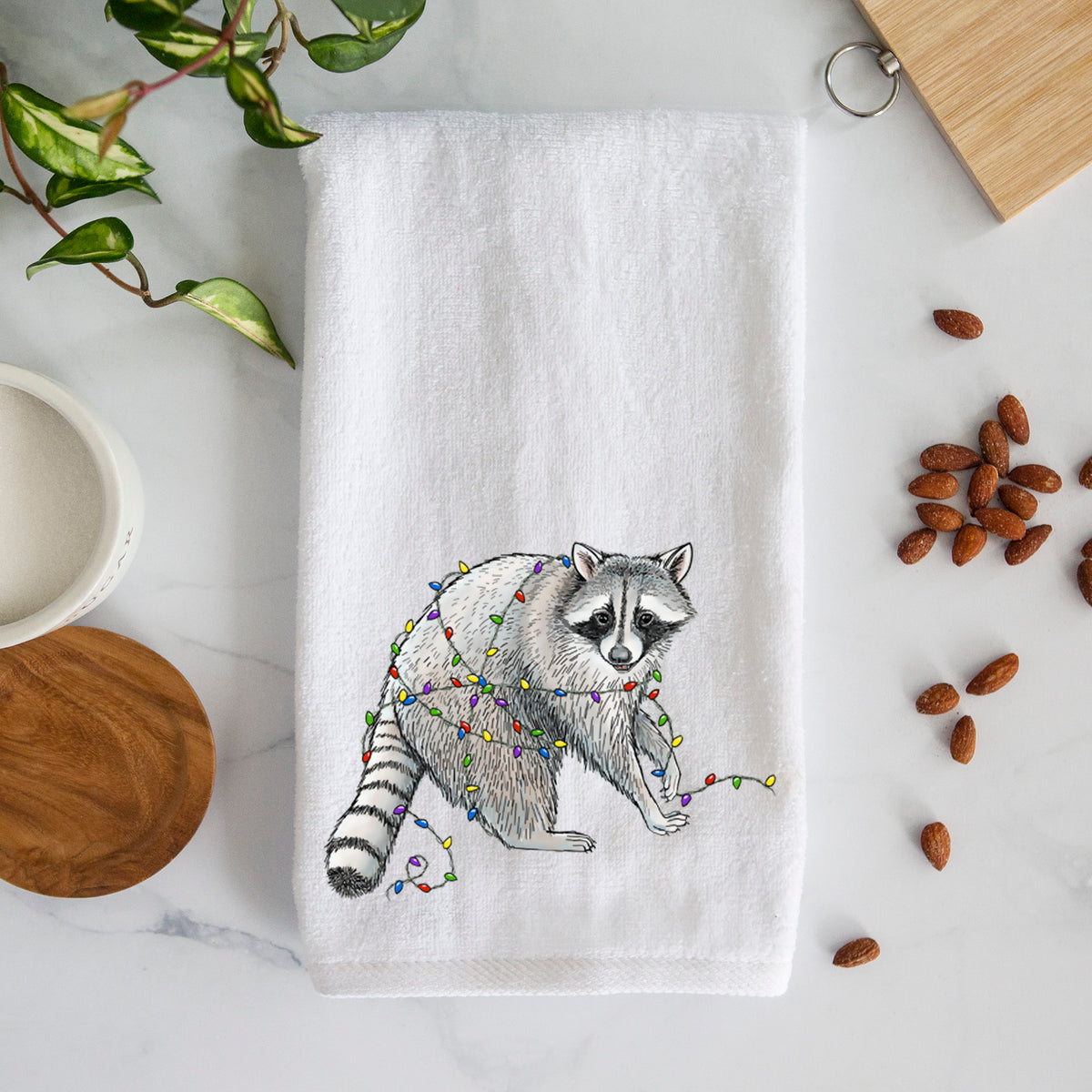 White hand towel printed with a black ink raccoon tangled in a string of colorful holiday lights.