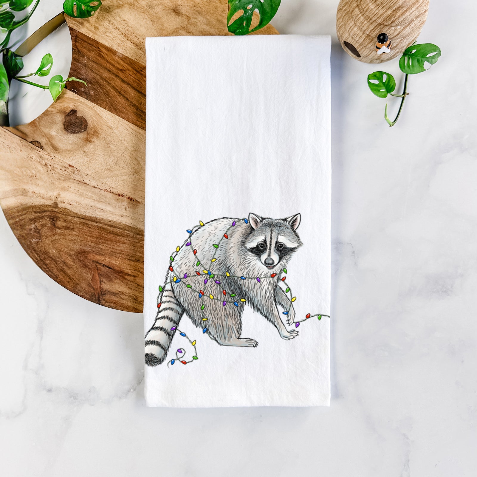 White cotton tea towel printed with a black ink raccoon tangled in a string of colorful holiday lights, on a marble counter beside a round wooden cutting board and greenery.