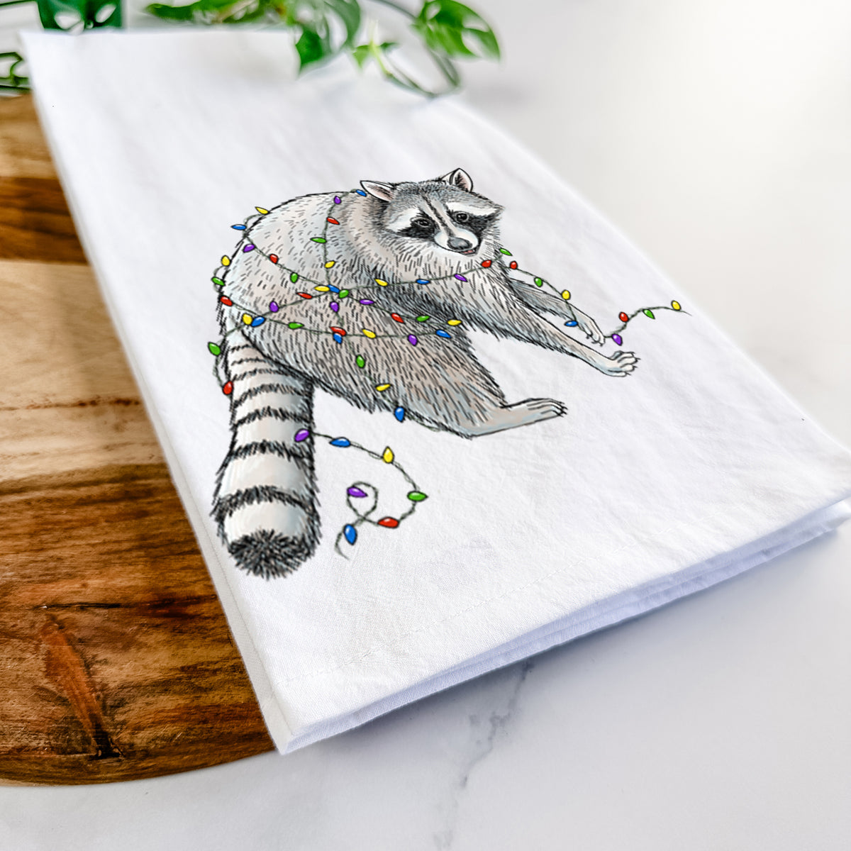 Close-up of a folded white tea towel on a wooden board, showing a black ink raccoon tangled in a string of colorful holiday lights.