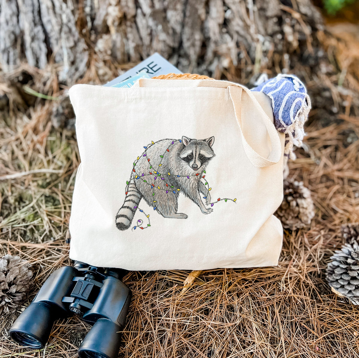 Canvas tote bag printed with a black ink raccoon tangled in a string of colorful holiday lights, resting on dry grass beside a pair of binoculars.