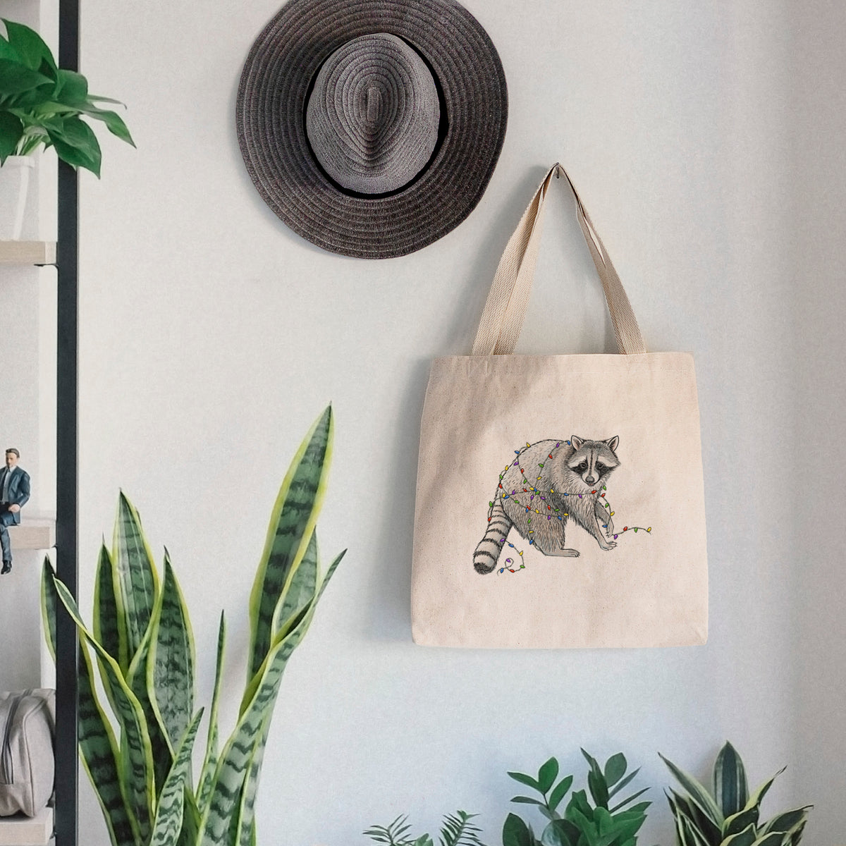 Canvas tote bag printed with a black ink raccoon tangled in a string of colorful holiday lights, hanging on a wall hook below a hat, next to potted plants.