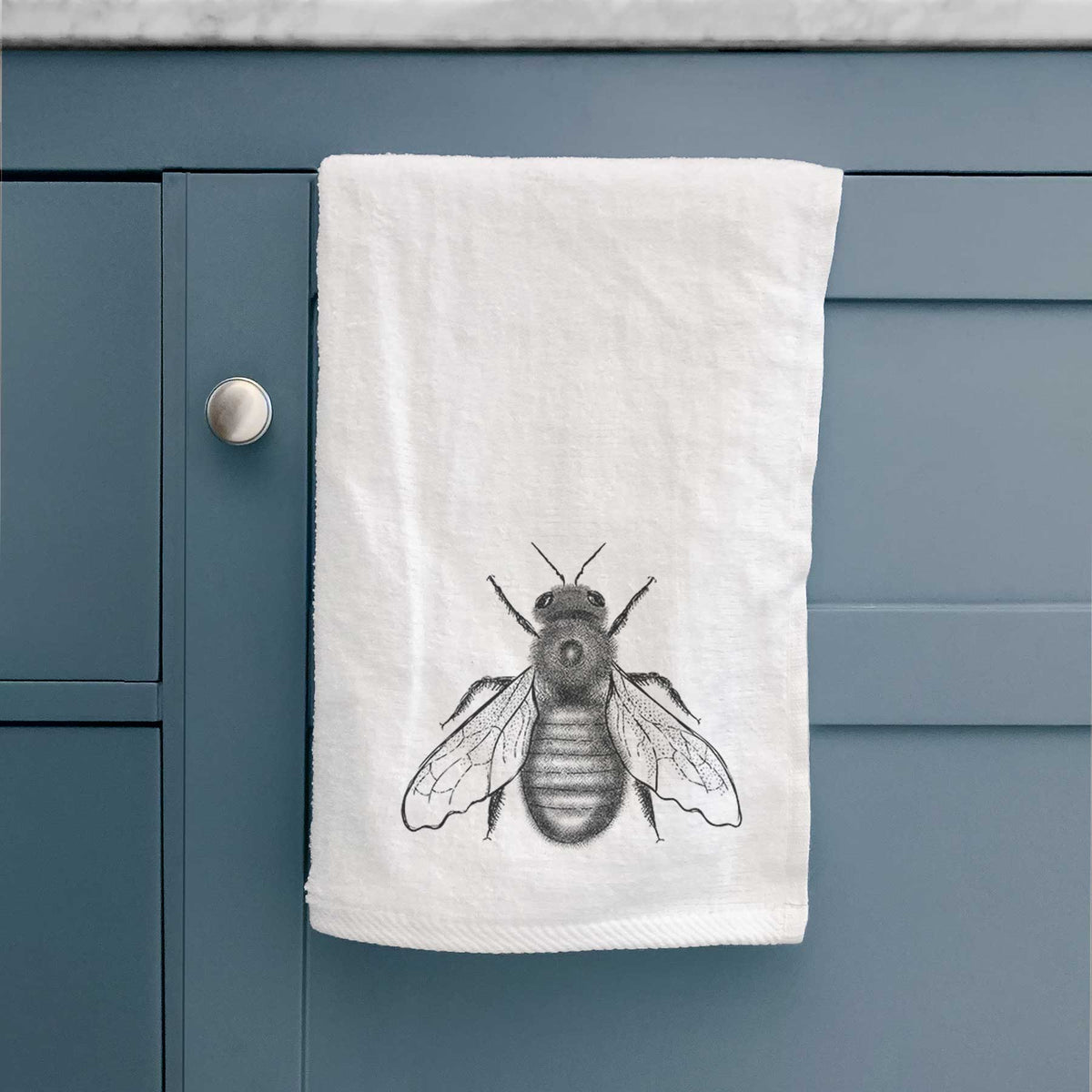 White hand towel printed with a detailed black ink illustration of an eastern carpenter bee with its wings spread, seen from above, hanging on a cabinet door.